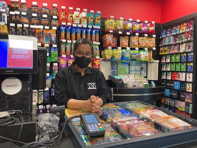 A store clerk stands behind a counter in a convenience shop, with lottery tickets and drinks visible.